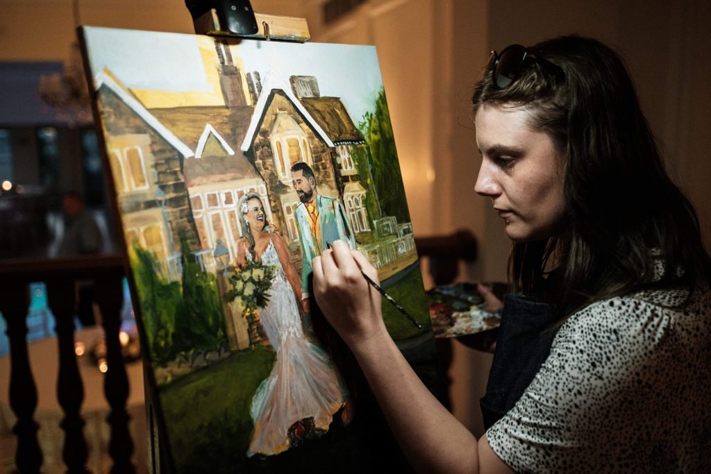 Behind-the-scenes photo of Katie Bowden painting a live wedding canvas outdoors, with views of the wedding venue’s crazy golf area and surrounding fields. This image highlights the relaxed outdoor setting captured through live painting.