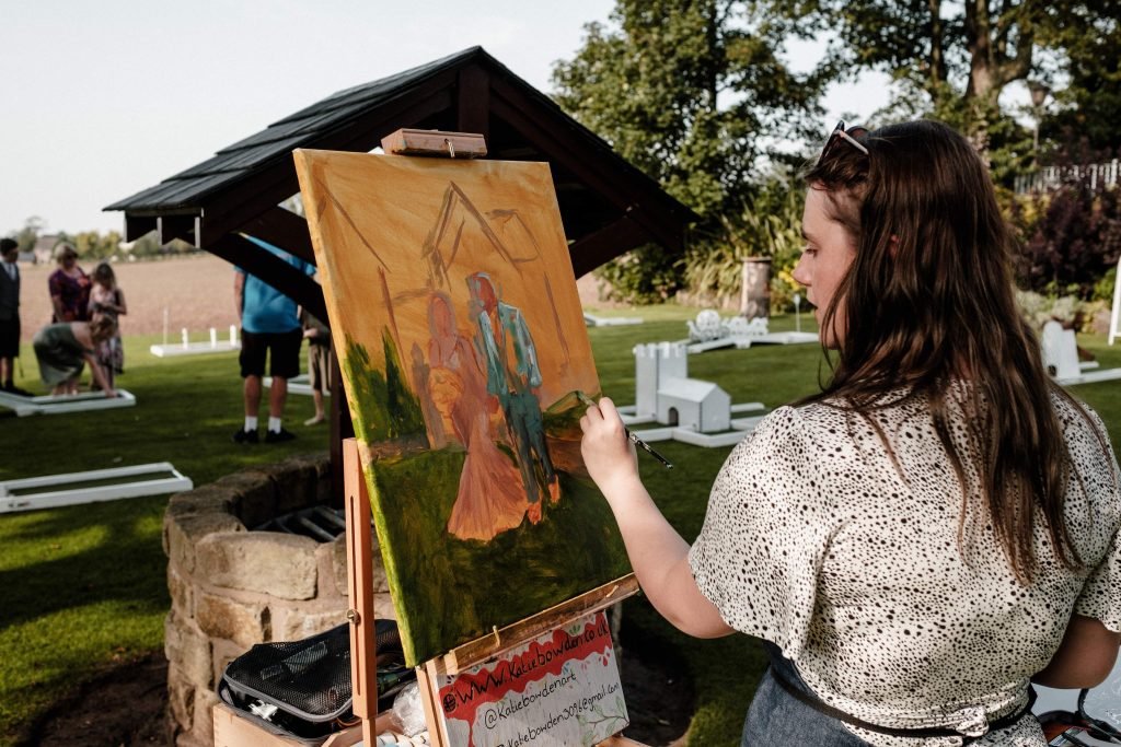 Katie Bowden painting a live wedding canvas outdoors with crazy golf and fields in the background
